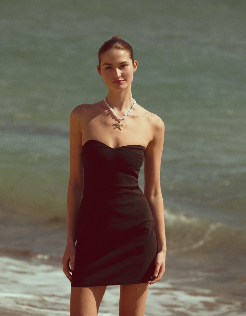 Bershka's summer 2024 editorial highlights Angelina Frerk in a chic black strapless dress by the beach.