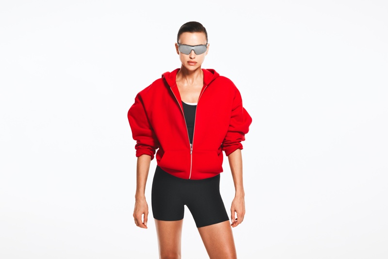 Irina Shayk poses in H&M Move's red hoodie and bike shorts with sunglasses.