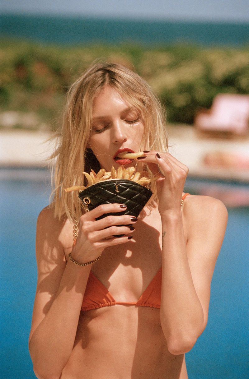 Anja Rubik eats French fries with Saint Laurent's Sade triangle coin purse from its Tan Lines capsule.