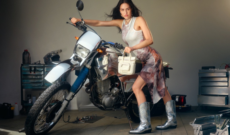 Wearing a printed dress and cowboy boots, Amelia Grey poses alongside a motorcycle for Desigual fall 2024 campaign.